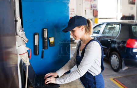 Photo for Female auto typing on laptop, ordering parts, communicating with customer. Beautiful woman working in a garage, wearing blue coveralls. Female automotive service technican. - Royalty Free Image