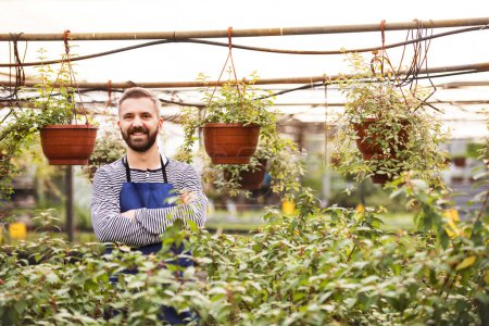 Photo for Portrait of gardener standing in the middle of plants, flowers and seedlings. Small greenhouse business. Offering wide range of plants during spring gardening season. - Royalty Free Image