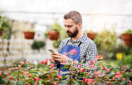 Gardener, business owner making phone call with customer or supplier, standing in front flowers and seedlings. Small greenhouse business.