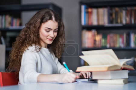 Photo for Young female student in library, focusing on final project, presentation. University student preparing for final exam, writing. - Royalty Free Image