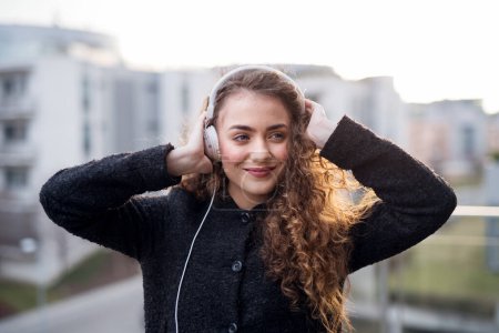 Hermosa mujer con el pelo rizado de pie en la terraza, disfrutando de la fría mañana de primavera, escuchando música clamando, afirmaciones positivas, usando auriculares.