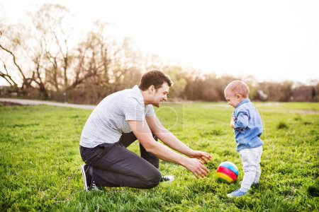 Foto de Padre jugando con el niño pequeño en la hierba, divirtiéndose durante el cálido día de primavera. Concepto del día de los padres. Tiempo en familia al aire libre - Imagen libre de derechos