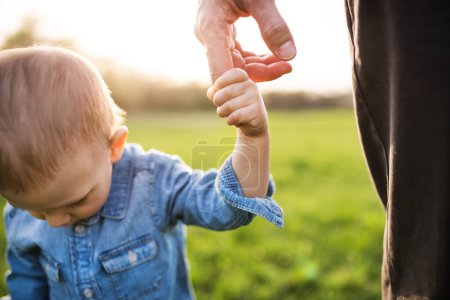 Foto de Retrato del padre sosteniendo al niño pequeño con el dedo, caminando en la naturaleza primaveral. Primeros pasos para el niño durante el cálido día de primavera. Concepto del día de los padres. De cerca. - Imagen libre de derechos