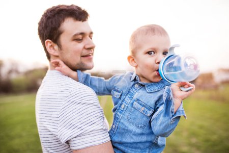 Foto de Un padre sosteniendo a un niño pequeño mientras bebe agua de la botella. Cálido día de primavera al aire libre. Concepto del día de los padres. - Imagen libre de derechos