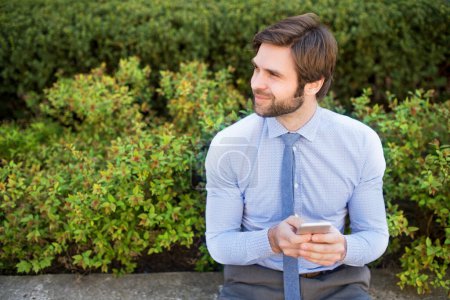Téléchargez les photos : Bel homme d'affaires tenant smartphone assis sur un banc dans le parc de la ville. Je travaille à distance, j'attends une réunion d'affaires. Gestionnaire souriant, extérieur en milieu urbain - en image libre de droit
