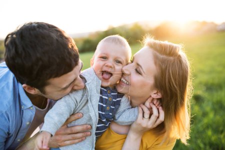 Foto de Nuevos padres sosteniendo pequeño niño, bebé, al aire libre en la naturaleza de primavera. Haciendo reír al bebé, besándolo en las mejillas. - Imagen libre de derechos