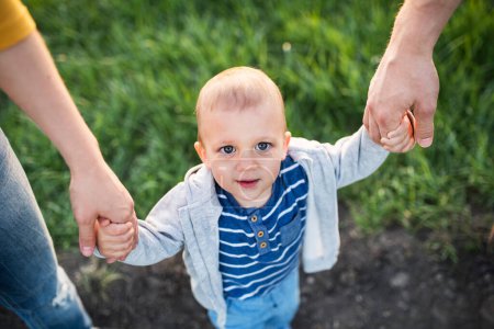 Foto de Nuevos padres sosteniendo a un niño pequeño, con las manos, balanceándolo. Familia al aire libre a pie en la naturaleza de primavera. Feliz momento familiar. - Imagen libre de derechos