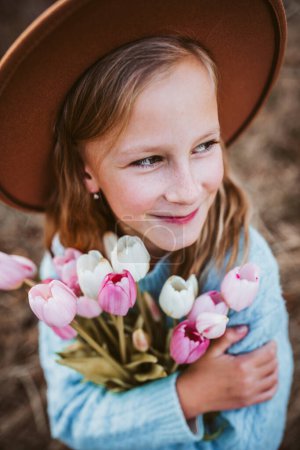 Foto de Retrato de una hermosa chica con sombrero, sosteniendo un ramo de tulipanes rosados. Momento de primavera, infancia en la naturaleza. - Imagen libre de derechos
