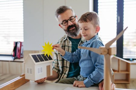 Photo for Father explaining renewable green energy, teaching about sustainable lifestyle his young son. Playing with model of house with solar panels, wind turibine at home. Learning through play. - Royalty Free Image