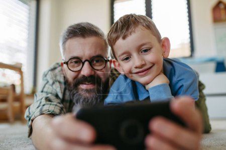 Téléchargez les photos : Petit garçon faisant selfie sur smartphone avec son père, couché sur le sol dans la chambre des enfants, faire des visages stupides. Papa expliquant la technologie au fils, littératie numérique pour les enfants. - en image libre de droit