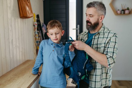 Father helping son get ready for a kindergarten, preschool. Putting backpack on his back.