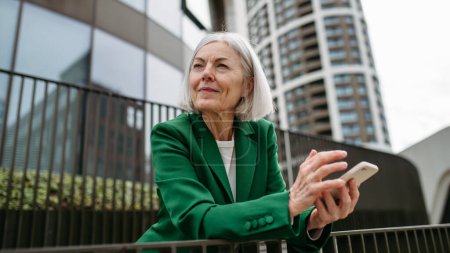 Reife Geschäftsfrau hält Smartphone in der Hand, wartet auf Geschäftspartner in der Stadt Schöne ältere Frau mit grauen Haaren steht auf der Straße der Stadt und lächelt.
