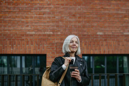 Téléchargez les photos : Portrait de femme mature élégante avec des cheveux gris sur la rue de la ville, en plein air. Femme plus âgée veste en cuir avec sourire doux sur le visage. - en image libre de droit