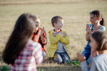 Photo for Children sitting on grass on meadow playing clapping game. Dedicated teachers during outdoor active education teaching about ecosystem, ecology and nature. - Royalty Free Image