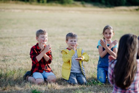 Photo for Children sitting on grass on meadow playing clapping game. Dedicated teachers during outdoor active education teaching about ecosystem, ecology and nature. - Royalty Free Image