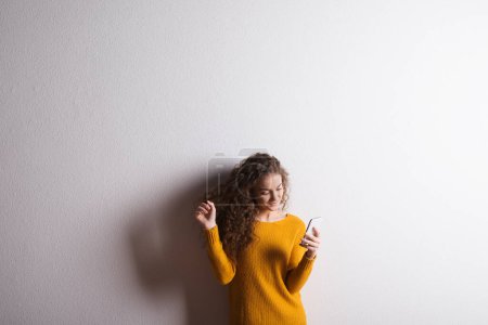 Photo for Portrait of a gorgeous teenage girl with curly hair, scrolling on smartphone. Studio shot, white background with copy space. - Royalty Free Image