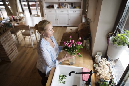 Beautiful mature woman arranging a bouquet of roses in a vase, a hobby and relaxation. Older woman living alone, enjoying peaceful weekend day.