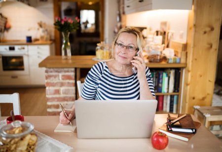 Older woman working from homeoffice, typing on laptop in the kitchen and making phone calls. The retiree earning extra money during retirement.