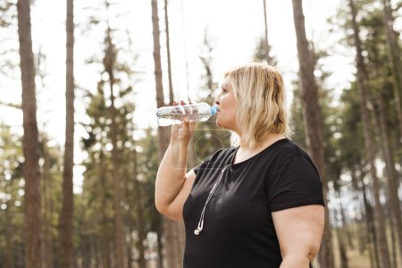 Téléchargez les photos : Femme en surpoids boire de l'eau après une course dans la nature. Exercice en plein air pour les personnes obèses - en image libre de droit
