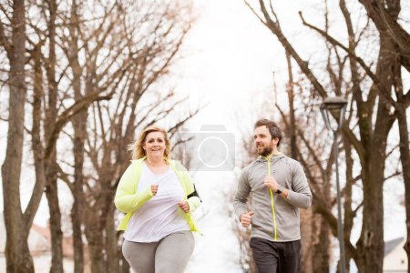 Overweight woman running in city park, personal trainer checking her performance. Exercising outdoors for people with obesity, support from friend or fitness coach.