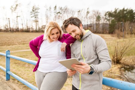 Overweight woman resting after run, personal trainer checking her profile, performance on tablet. Exercising outdoors for people with obesity, support from male fitness coach.