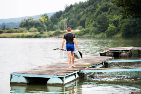 El joven piragüista lleva canoa y remo, va al agua, camina sobre un muelle de madera. Concepto de piragüismo como deporte dinámico y aventurero.