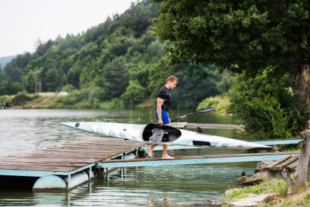 Young canoeist carry canoe and paddle, going into water, walking on wooden dock. Concept of canoeing as dynamic and adventurous sport.