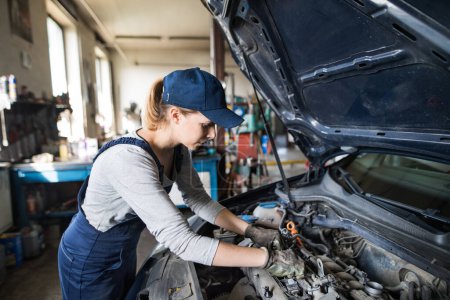 Photo for Female auto mechanic repairing, maintaining a car. Beautiful woman working in a garage, wearing blue coveralls. Female automotive service technican. - Royalty Free Image