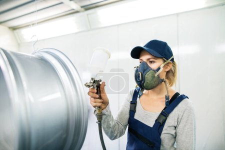 Photo for Female car painter, paingting vehicles in auto body shop. Young woman holding spray gun, spraying paint on rim of wheel, wearing mask and coveralls. - Royalty Free Image