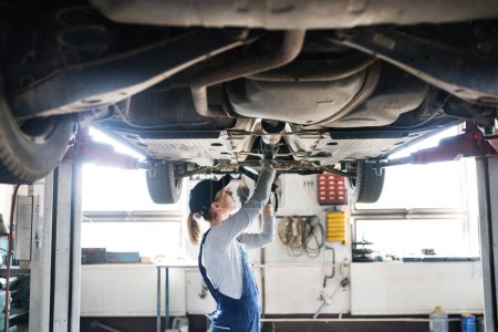 Photo for Female auto mechanic elevating car on car, automotive lift, working underneath. Beautiful woman working in a garage, wearing blue coveralls. Female automotive service technican. - Royalty Free Image