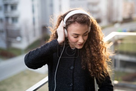 Beautiful woman with curly hair standing on terrace, enjoying cold spring morning, listening claming music, positive affirmations, wearing headphones.