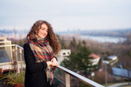 Hermosa mujer con el pelo rizado de pie en la terraza, disfrutando de la fría mañana de otoño, día soleado. Disfrutando de la vista, sintiéndose mentalmente mejor. Hablando afirmaciones positivas.