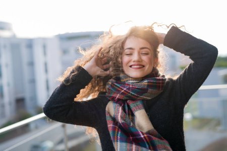 Schöne Frau mit lockigem Haar steht auf der Terrasse und genießt den kalten Herbstmorgen, den sonnigen Tag. Aussicht genießen, sich geistig besser fühlen. Positive Bejahungen.