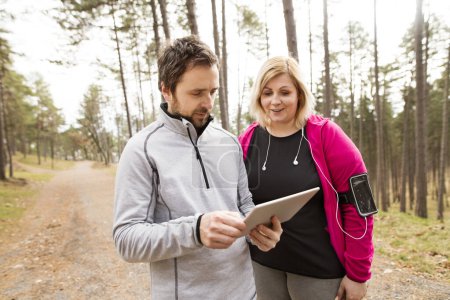 Overweight woman resting after run, personal trainer checking her profile, performance on tablet. Exercising outdoors for people with obesity, support from male fitness coach.