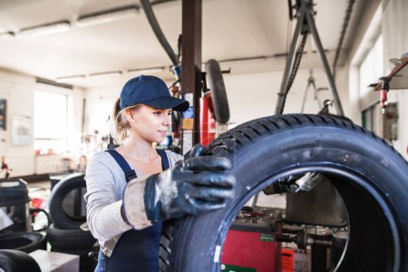 Photo for Female auto mechanic changing tieres in auto service. Beautiful woman holding tire in a garage, wearing blue coveralls. Tire mounting. Female automotive service technican. - Royalty Free Image