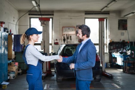 Female auto mechanic talking with customer, shaking hands. Beautiful woman working in a garage, wearing blue coveralls. Female automotive service technican.