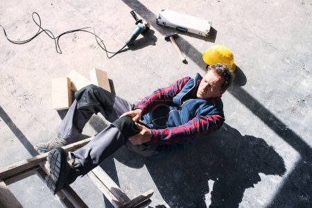 Male worker lying on the floor after fall from ladder, holding his knee. Work injury, accident in workplace. Work injury, accident in workplace.