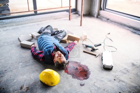Foto de Un trabajador inconsciente tirado en el suelo después de un accidente en la obra, sangre en la cabeza. Lesiones laborales, accidentes de trabajo. - Imagen libre de derechos