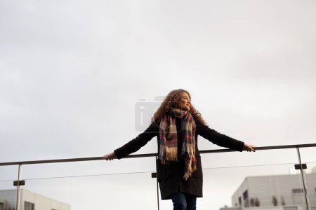 Foto de Hermosa mujer con el pelo rizado de pie en la terraza, disfrutando de la fría mañana de otoño, día soleado. Disfrutando de la vista, sintiéndose mentalmente mejor. Hablando afirmaciones positivas. - Imagen libre de derechos
