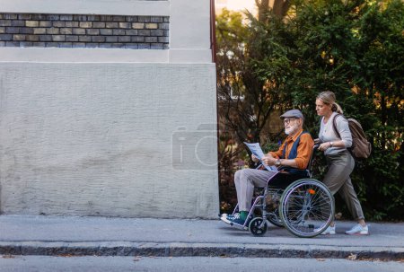 Enkelin schubst Senior im Rollstuhl auf Straße Zeitungskauf am Kiosk. Pflegerin und älterer Mann genießen einen warmen Herbsttag auf dem Heimweg von einem Einkaufsbummel.