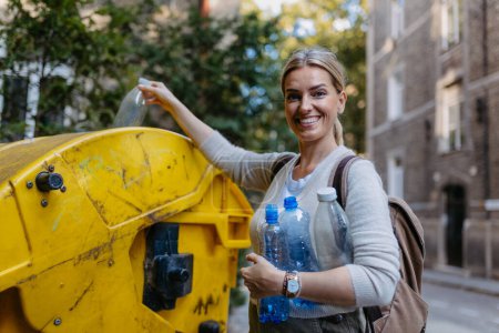 Frau wirft Plastikmüll, Flaschen in Recyclingcontainer vor ihrer Wohnanlage. Frau sortiert den Abfall nach Material in farbige Tonnen.