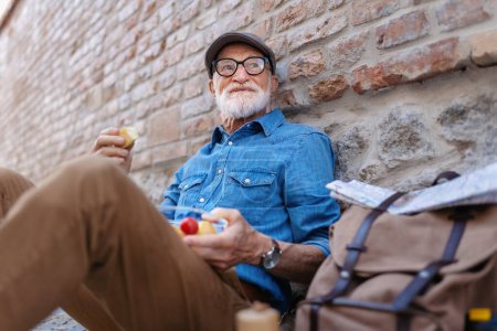 Foto de Anciano con mochila sentado junto a la pared de ladrillo, tomando bocadillos caseros saludables y descansando. Viajes y viajes en solitario durante la jubilación. - Imagen libre de derechos