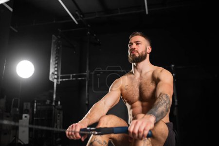 Hombre haciendo ejercicio en la máquina de remo, usando solo pantalones cortos, pecho desnudo. Entrenamiento de rutina para una salud física y mental.