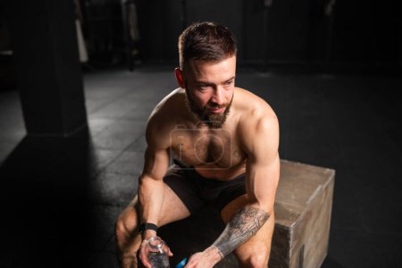Muskulöser Mann, der sich nach dem Sport ausruht, Wasser aus der Flasche trinkt, sitzt und eine kurze Hose mit muskulösem nacktem Oberkörper trägt. Routinetraining für körperliche und geistige Gesundheit.