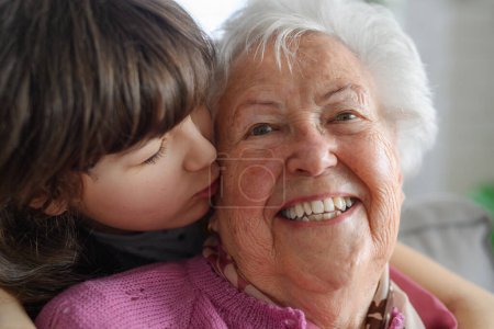 Foto de Linda chica abrazando y besando gradmother en su mejilla por detrás. Retrato de una anciana que pasa tiempo con su nieta. - Imagen libre de derechos