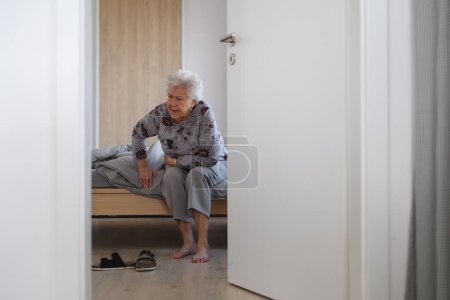 Senior woman trying to stand up from bed, feeling pain, holding abdomen.