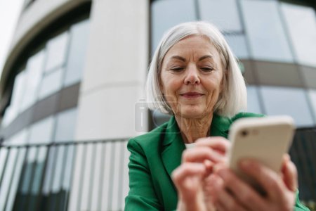 Téléchargez les photos : Femme d'affaires mature tenant smartphone, attendant un partenaire d'affaires dans la villeBelle femme plus âgée avec les cheveux gris debout sur la rue de la ville, souriant. - en image libre de droit