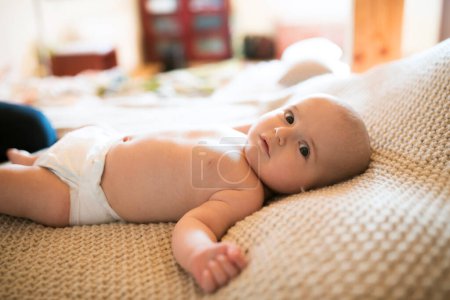 Portrait of cute little baby boy in diaper lying on bed, on back, looking at camera.