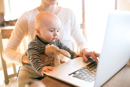 Mutter mit kleinem Baby arbeitet vom Homeoffice aus und tippt auf dem Laptop. Mutter bezahlt Rechnungen online und hält kleines Baby in der Hand.