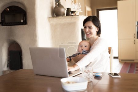 Mutter mit kleinem Baby arbeitet vom Homeoffice aus und tippt auf dem Laptop. Mutter bezahlt Rechnungen online und hält kleines Baby in der Hand.
